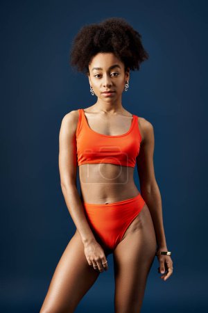 Foto de Mujer afroamericana con estilo alcanza una pose en bikini naranja vibrante sobre un audaz telón de fondo azul. - Imagen libre de derechos