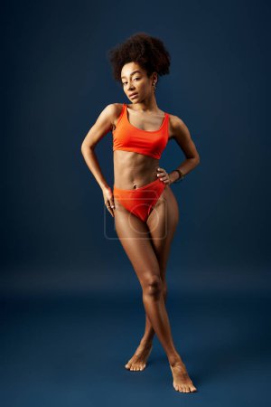 Foto de A stylish, young African American woman poses in an orange bikini against a blue background. - Imagen libre de derechos