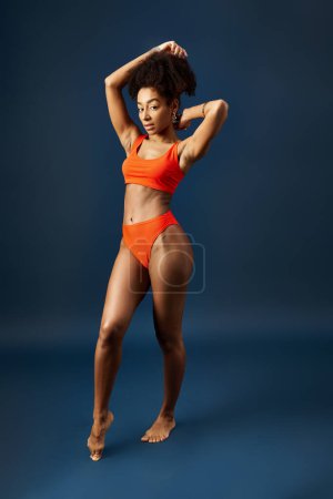 Elegante mujer afroamericana en bikini naranja posando sobre un fondo azul brillante.