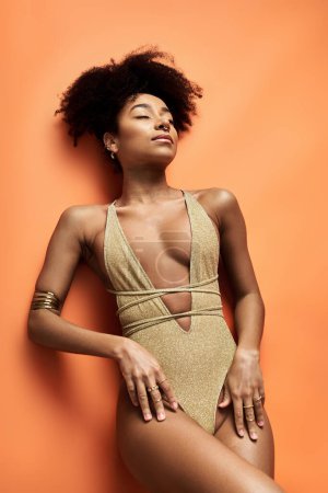 Téléchargez les photos : Femme afro-américaine élégante en maillot de bain en or frappe une pose sur fond orange vif. - en image libre de droit