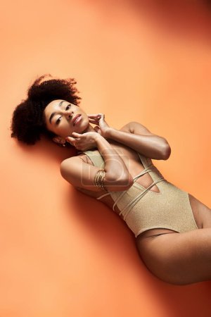 Stylish African American woman sunbathing in trendy swimsuit on bright orange background.