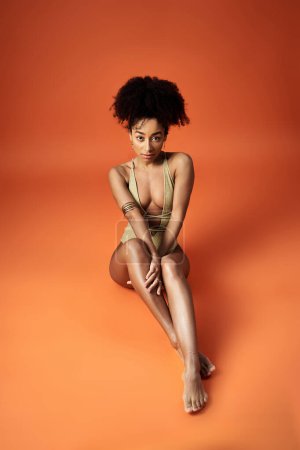 Photo for Stylish African American woman in trendy swimsuit seated on vibrant orange backdrop. - Royalty Free Image
