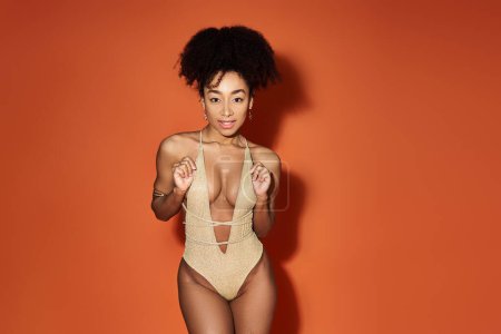 Photo for A captivating young African American woman in a trendy gold swimsuit striking a pose against a bright orange backdrop. - Royalty Free Image