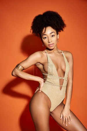 Stylish young African American woman posing in tan swimsuit on orange background.