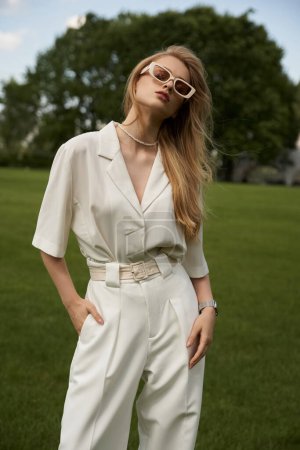 A stylish woman in a white jumpsuit strikes a pose in a vast, green field surrounded by serenity and beauty.