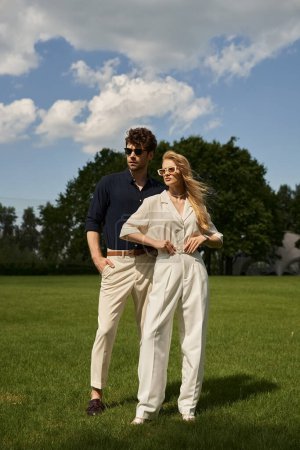 A stylish man and woman in elegant attire stand in a lush field, surrounded by the vibrant greenery of nature.
