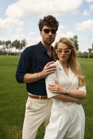 A stylish man and woman strike a pose in an expansive green field, exuding elegance and sophistication.