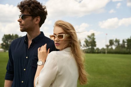 A stylish couple, donning sunglasses, stands together in a lush field, embodying sophisticated flair and a touch of old-world charm.