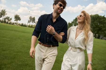 Téléchargez les photos : A beautiful young couple dressed in elegant clothing enjoys a leisurely walk in the park on a green field, embodying old money style and rich people lifestyle. - en image libre de droit