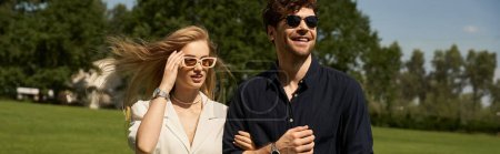 Ein glamouröser junger Mann und eine junge Frau in stylischer Kleidung spazieren durch ein üppiges Feld und tragen trendige Sonnenbrillen unter der Sonne.