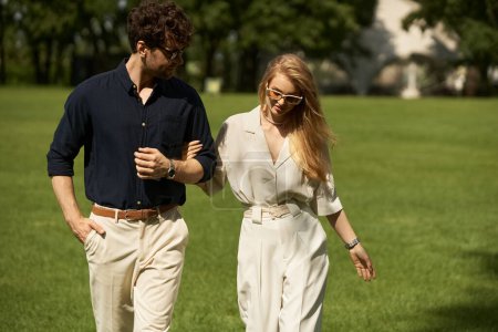 A stylish young couple, elegantly dressed, leisurely walk together in a lush park setting.