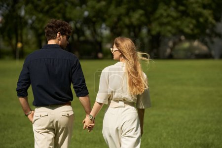 Foto de Una hermosa pareja joven con elegante atuendo tomados de la mano mientras camina por un parque en un día soleado. - Imagen libre de derechos