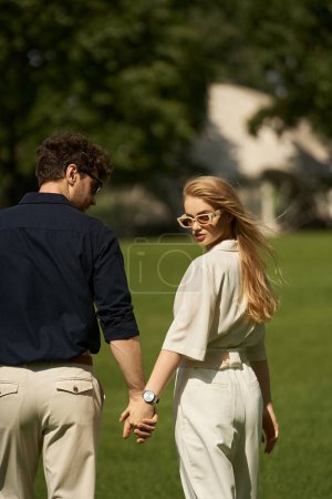 Foto de Una joven pareja elegantemente vestida, cogida de la mano en un parque, rodeada de exuberante vegetación, encarnando una sensación de elegancia atemporal. - Imagen libre de derechos