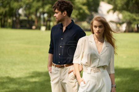 A beautiful, stylish young couple in elegant clothing walking together in a lush green park filled with vibrant foliage.