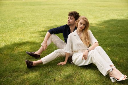 A classy young couple in elegant attire relax on the lush green grass, basking in each others company