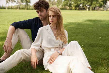 A stylish, young man and woman in elegant attire sit closely together on the lush green grass.