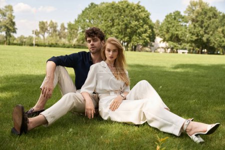 Téléchargez les photos : Couple élégant en tenue raffinée profitant d'une journée tranquille assis sur l'herbe dans un parc luxuriant. - en image libre de droit