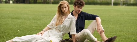 A stylish man and woman in elegant attire relax on the grass, enjoying each others company in a picturesque outdoor setting.