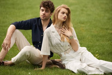 Ein junges Paar in eleganter Kleidung sitzt zusammen auf dem Gras und verkörpert einen luxuriösen Lebensstil in einer ruhigen Outdoor-Umgebung.