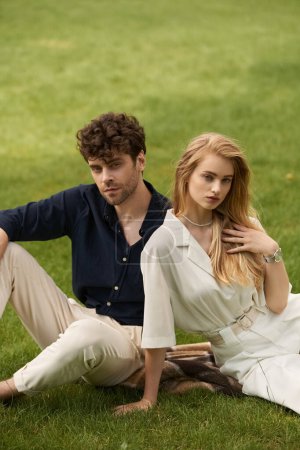 A stylish couple in elegant attire sits together on green grass, enjoying each others company in a serene outdoor setting.