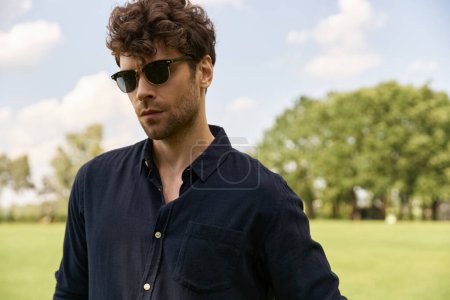 A stylish man in sunglasses stands confidently in a vast field, soaking in the tranquility around him.