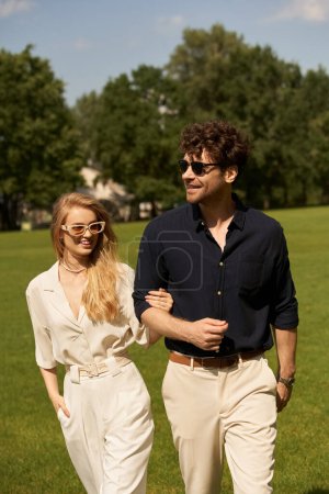 A young, elegant couple dressed in fine attire leisurely strolling through a lush green park on a sunny day.