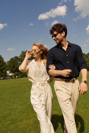 A stylish duo, dressed in elegant attire, leisurely walk through a verdant field.