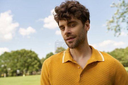 A stylish man in a vibrant yellow shirt stands amidst the lush greenery of a park on a sunny day.