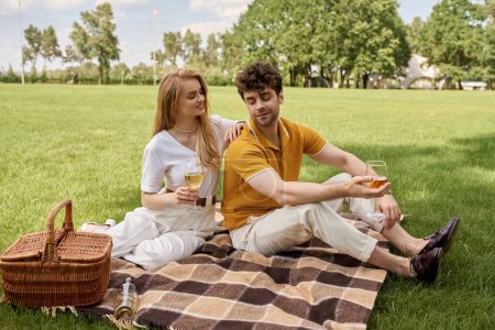Stilvolles junges Paar in eleganter Kleidung bei einem Picknick auf einem üppigen Parkfeld, das einen luxuriösen und raffinierten Lebensstil verkörpert.