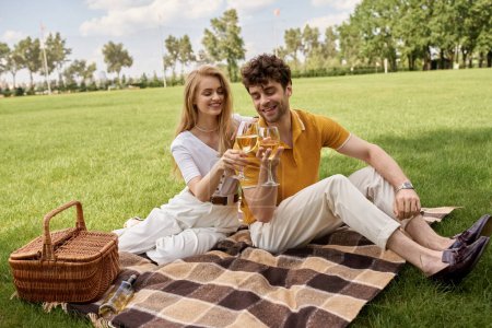 Un hombre y una mujer sofisticados en trajes elegantes sentados en una manta cerca de una cesta de picnic en un exuberante parque.