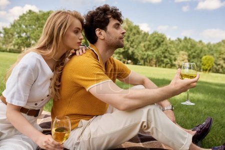 Stylish couple enjoying a romantic picnic in a park, holding wine glasses and immersed in conversation.