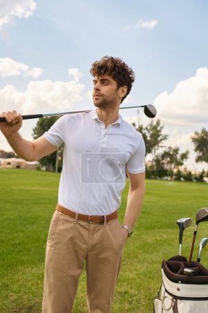 Ein Mann in eleganter Kleidung hält eine Golftasche und einen Golfschläger auf einer grünen Wiese in einem renommierten Golfclub.