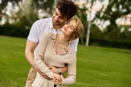 A young man and woman dressed in elegant attire hug affectionately in a serene park setting.