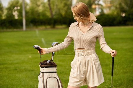Téléchargez les photos : Une jeune femme tient élégamment un sac de golf sur un terrain vert dans un club de golf, profitant d'une journée tranquille en plein air. - en image libre de droit