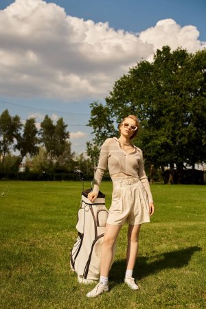 A sophisticated young woman stands gracefully in a field with a golf bag, enjoying the outdoors with an air of refined elegance.