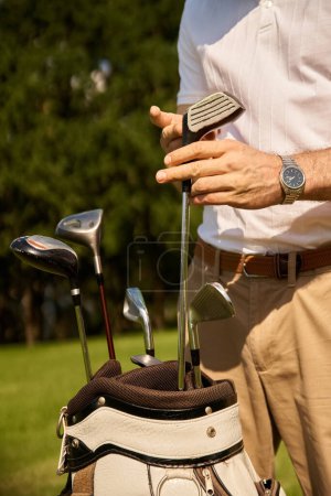 Ein stilvoller junger Mann in eleganter Kleidung, der seine Golfschläger in einer Tasche auf einer grünen Wiese in einem renommierten Golfclub verstaut.