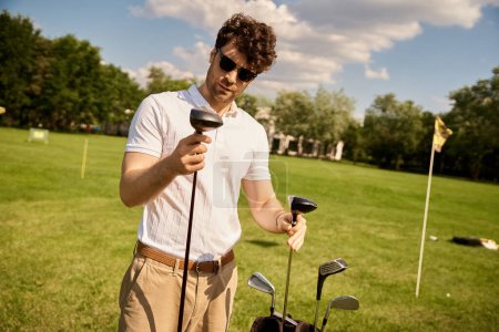 Téléchargez les photos : Homme élégant dans des vêtements élégants se tient à l'extérieur, tenant en toute confiance un club de golf dans ses mains sur un terrain vert luxuriant. - en image libre de droit
