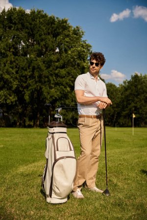 A man in casual attire stands by his golf bag on a lush golf course on a sunny day, enjoying a moment of relaxation.