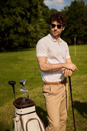 A man stands elegantly next to his golf bag on a green field at the golf club, embodying an old money style lifestyle.