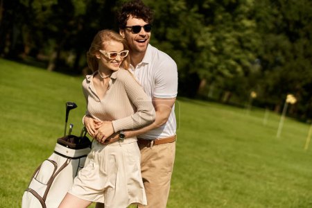 Foto de Un hombre joven y una mujer en traje elegante compartiendo un momento de afecto mientras posan en un campo de golf verde exuberante. - Imagen libre de derechos
