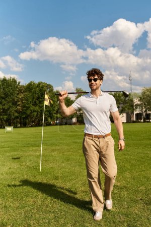 Ein junger Mann in eleganter Kleidung steht auf einer Rasenfläche und hält einen Golfschläger mit Eleganz und Stil in der Hand..