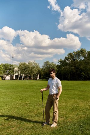 Ein stilvoller Mann steht auf einer grünen Wiese und ergreift einen Golfschläger, umgeben von friedlicher Schönheit der Natur.