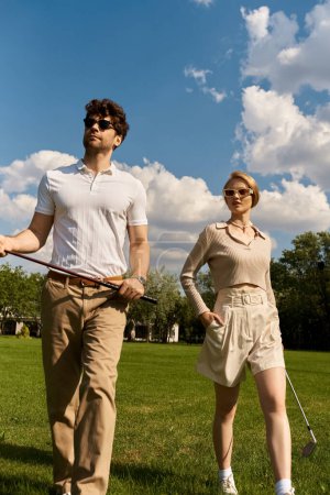 A stylish man and woman stroll across a vibrant field, holding golf clubs under the clear sky of a leisurely afternoon.