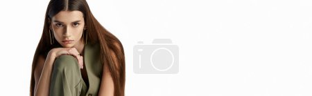 Téléchargez les photos : Une femme à la mode avec de longs cheveux bruns en tenue élégante, assise gracieusement sur un fond gris. - en image libre de droit