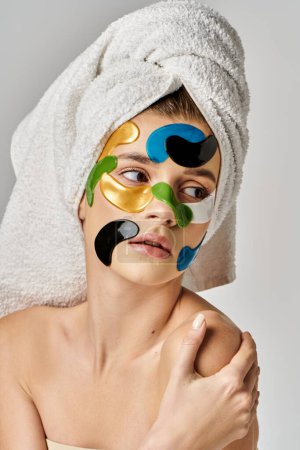 A serene young woman with a towel wrapped around her head with eye patches and makeup on.
