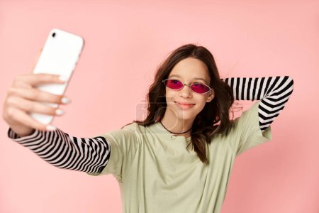 Stylish teenage girl in vibrant attire takes selfie with cellphone, wearing cool sunglasses.