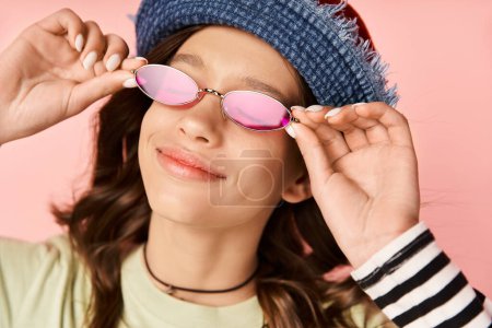 Foto de Una elegante adolescente posa en unas vibrantes gafas de sol rosadas y un sombrero azul de moda. - Imagen libre de derechos