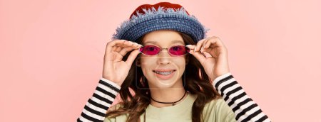 Ein stilvolles Teenager-Mädchen posiert selbstbewusst in lebendiger Kleidung, trägt einen trendigen Hut und eine Sonnenbrille.