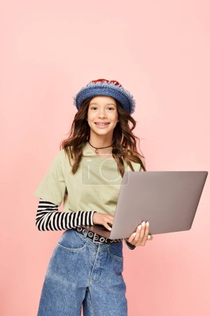 Téléchargez les photos : Adolescente élégante en jeans et un chapeau, tenant avec confiance un ordinateur portable. - en image libre de droit