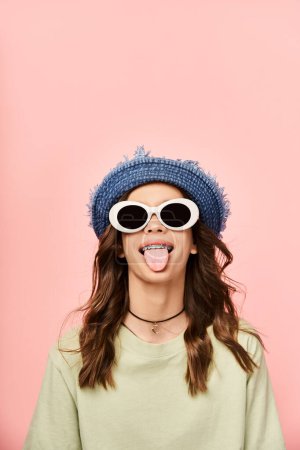 A stylish teenage girl in vibrant attire makes a funny face while wearing sunglasses and a hat.